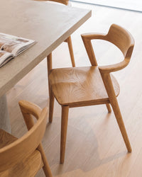 t4option0_1 | Jules dining chair in natural wood with curved armrests and a contoured seat, placed beside a dining table.