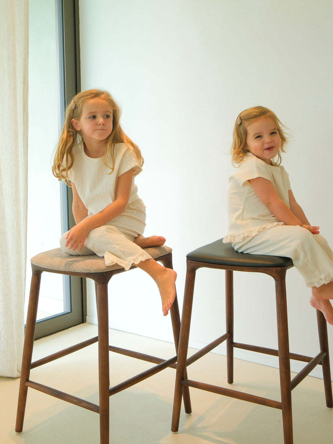 Two children sitting on Mowelli bar stools in walnut wood, one with fabric seat and one with leather seat, Belgian handcrafted furniture