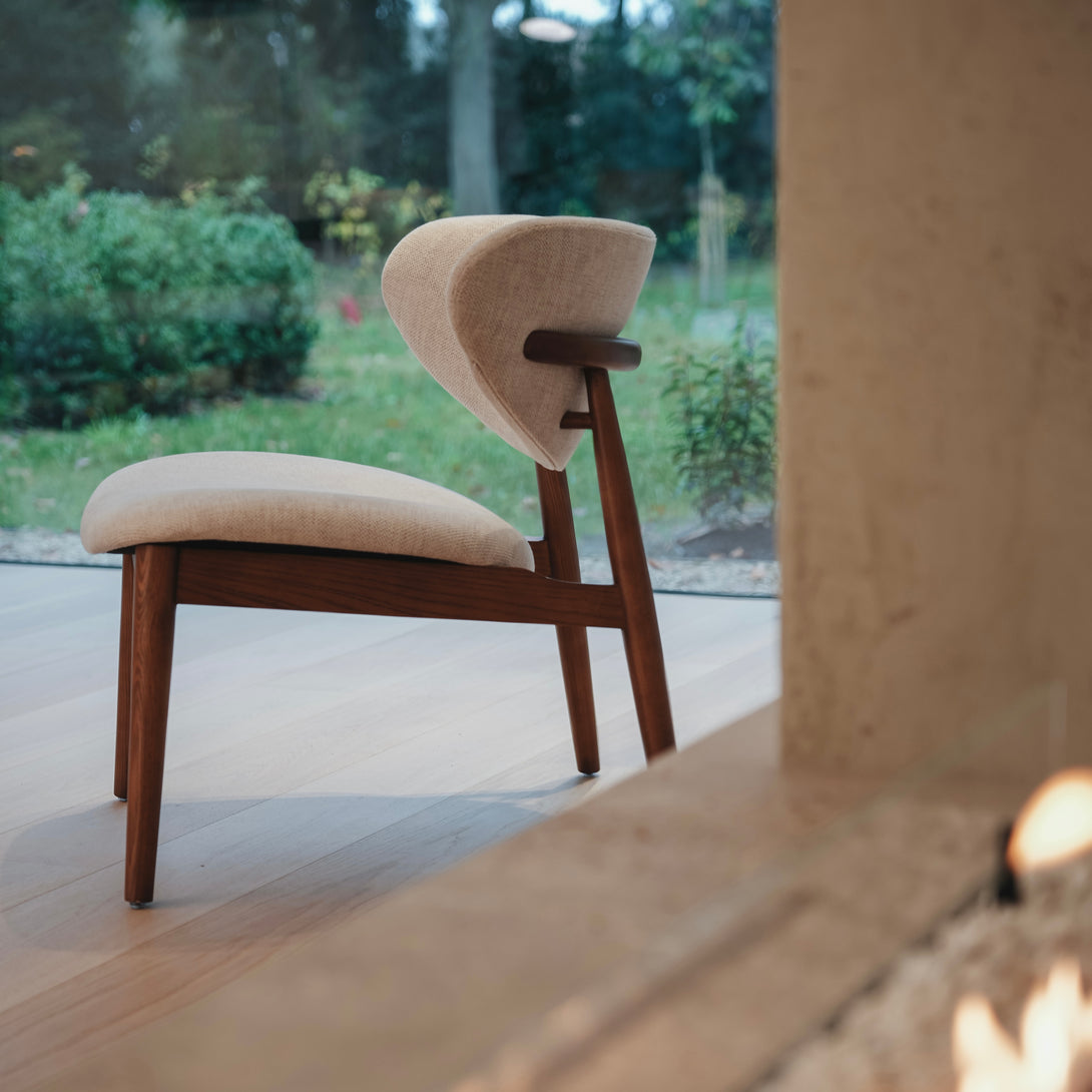 Side view of the Dianne lounge chair by Mowelli in walnut wood with beige upholstery, placed near a large window with a garden view.