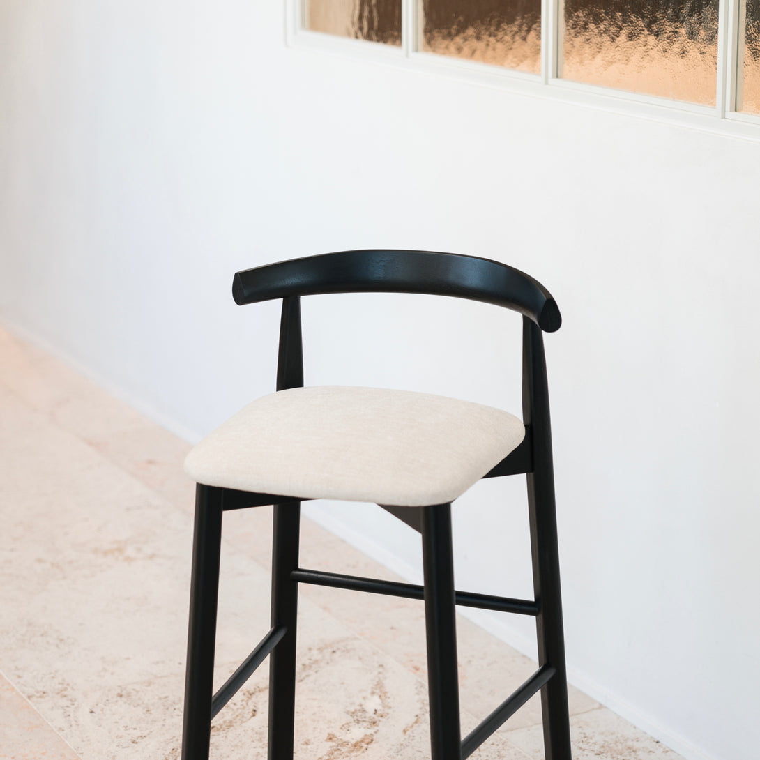 Isolated Ella bar stool by Mowelli in black wood and neutral fabric, displayed against a white wall.