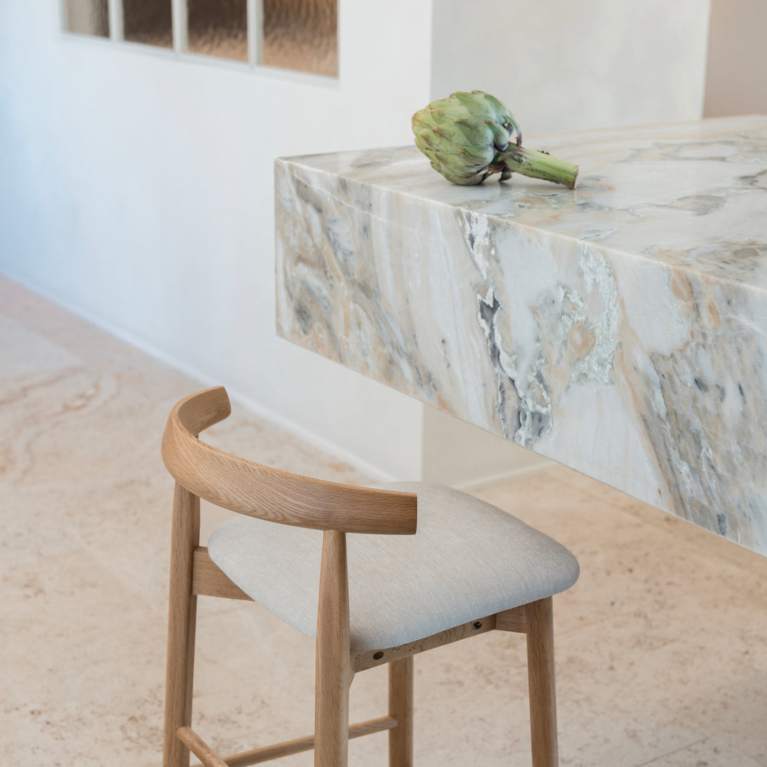 Natural oak Ella bar stool by Mowelli with light upholstered seat, placed at a marble kitchen island.