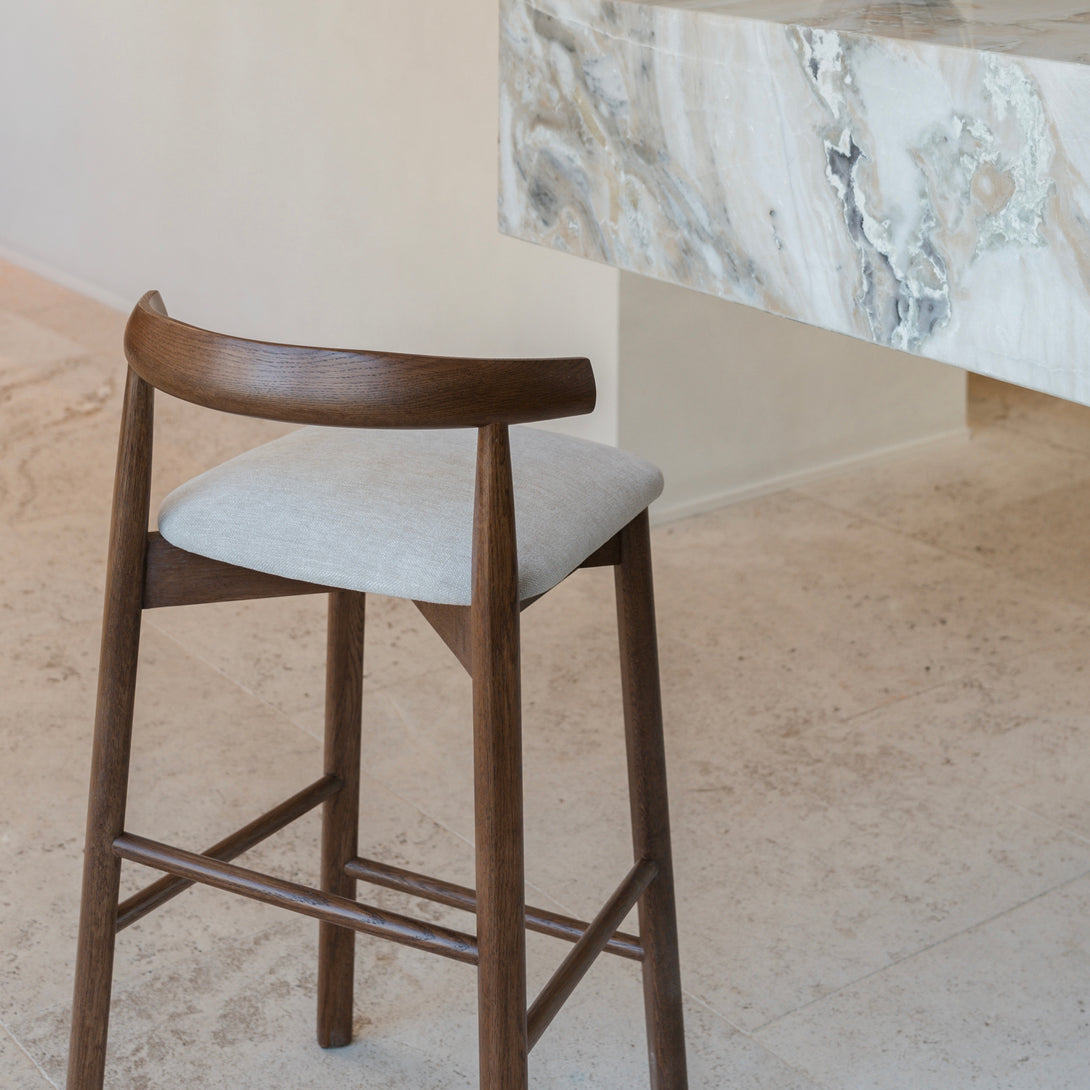 Back view of walnut Ella bar stool by Mowelli next to marble kitchen counter on travertine flooring.