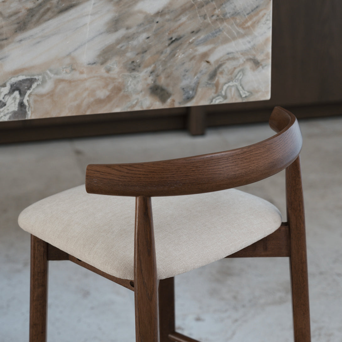 Back view of Mowelli's walnut Ella bar stool in front of marble kitchen counter, showing soft beige seating