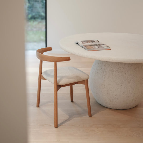 An elegant Ella dining chair by Mowelli with a upholstered seat and wooden backrest, placed next to a round table in a bright room.