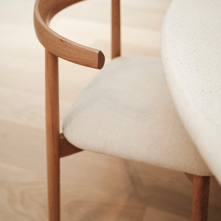 Close-up of Ella dining chair by Mowelli in light oak with soft beige upholstery, shown at a round dining table.