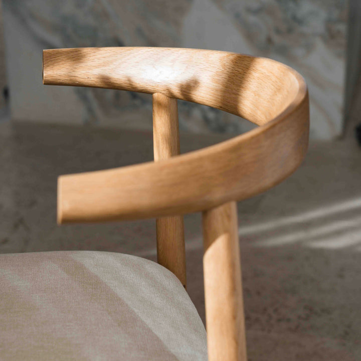 Close-up of natural oak Ella chair by Mowelli with soft fabric seat, sunlight casting gentle shadows on frame.