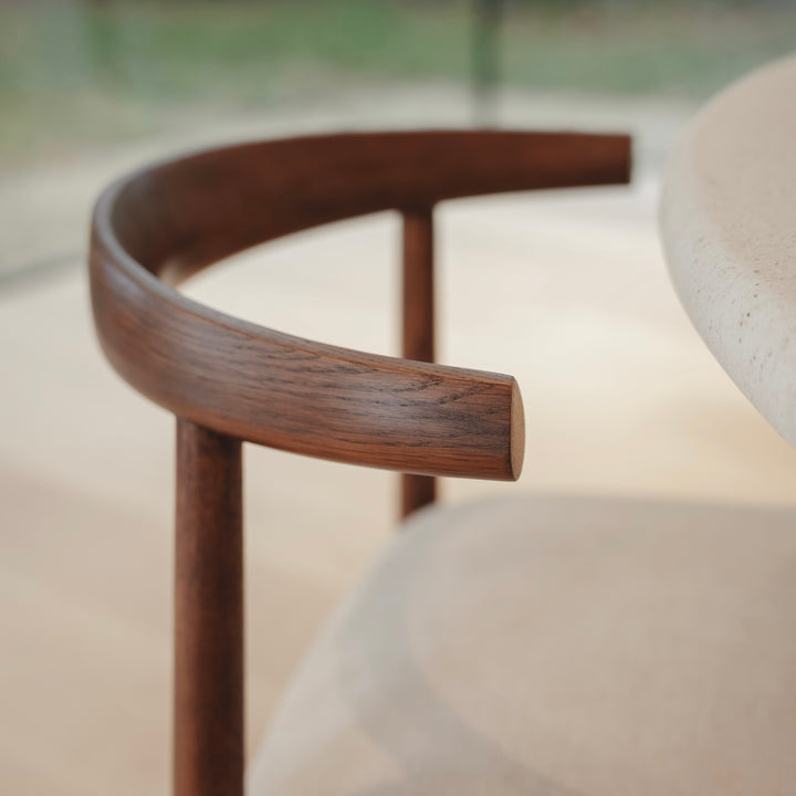 Close-up of Ella dining chair by Mowelli with a upholstered seat and wooden backrest, placed next to a round table in a bright room.