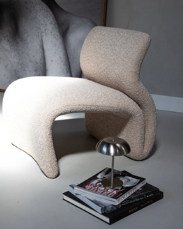 A sculptural, curved upholstered chair in a textured beige bouclé fabric, styled in a minimalist room setting with a small silver table lamp and books in the foreground.