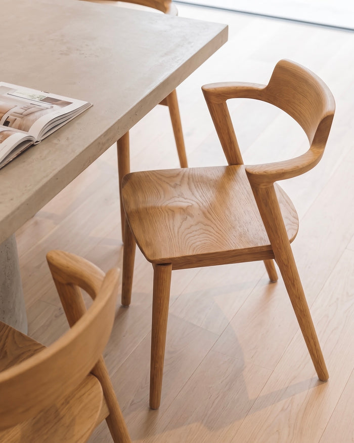 t4option0_1 | Jules dining chair in natural wood with curved armrests and a contoured seat, placed beside a dining table.