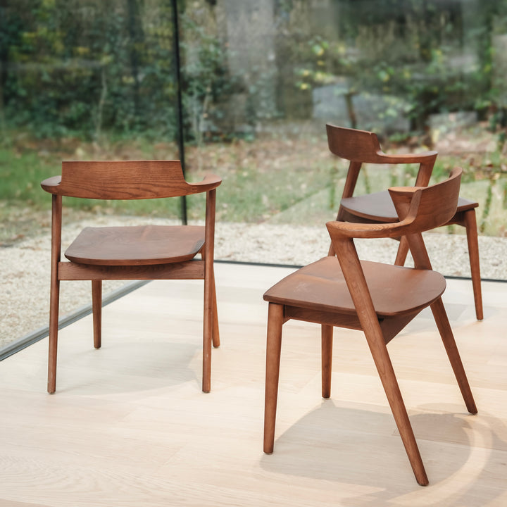 Set of Jules dining chairs by Mowelli standing before a window. Contemporary wooden chairs with curved armrests and Belgian craftsmanship