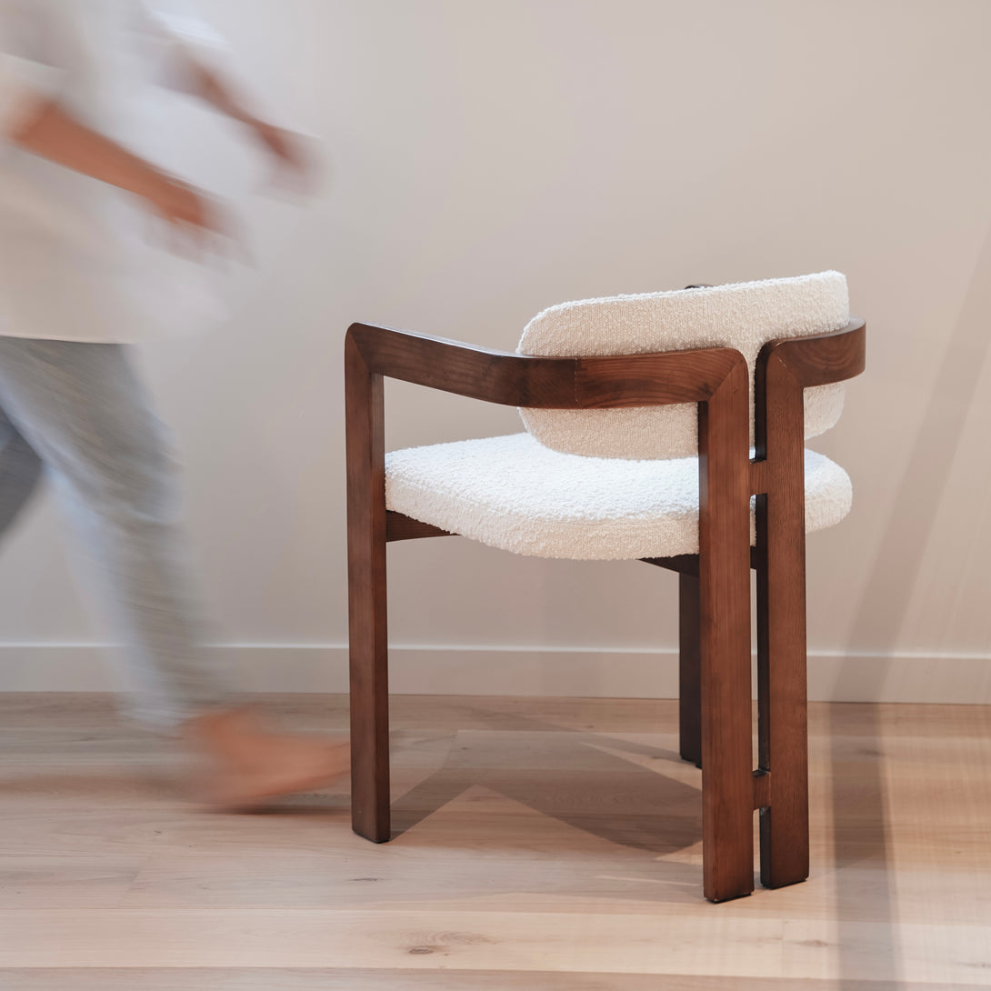 Mila chair by Mowelli with a walnut wood frame and white bouclé fabric seat and backrest, positioned on a light wood floor with a person walking in the background.