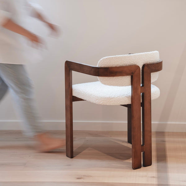 Mila chair by Mowelli with a walnut wood frame and white bouclé fabric seat and backrest, positioned on a light wood floor with a person walking in the background.