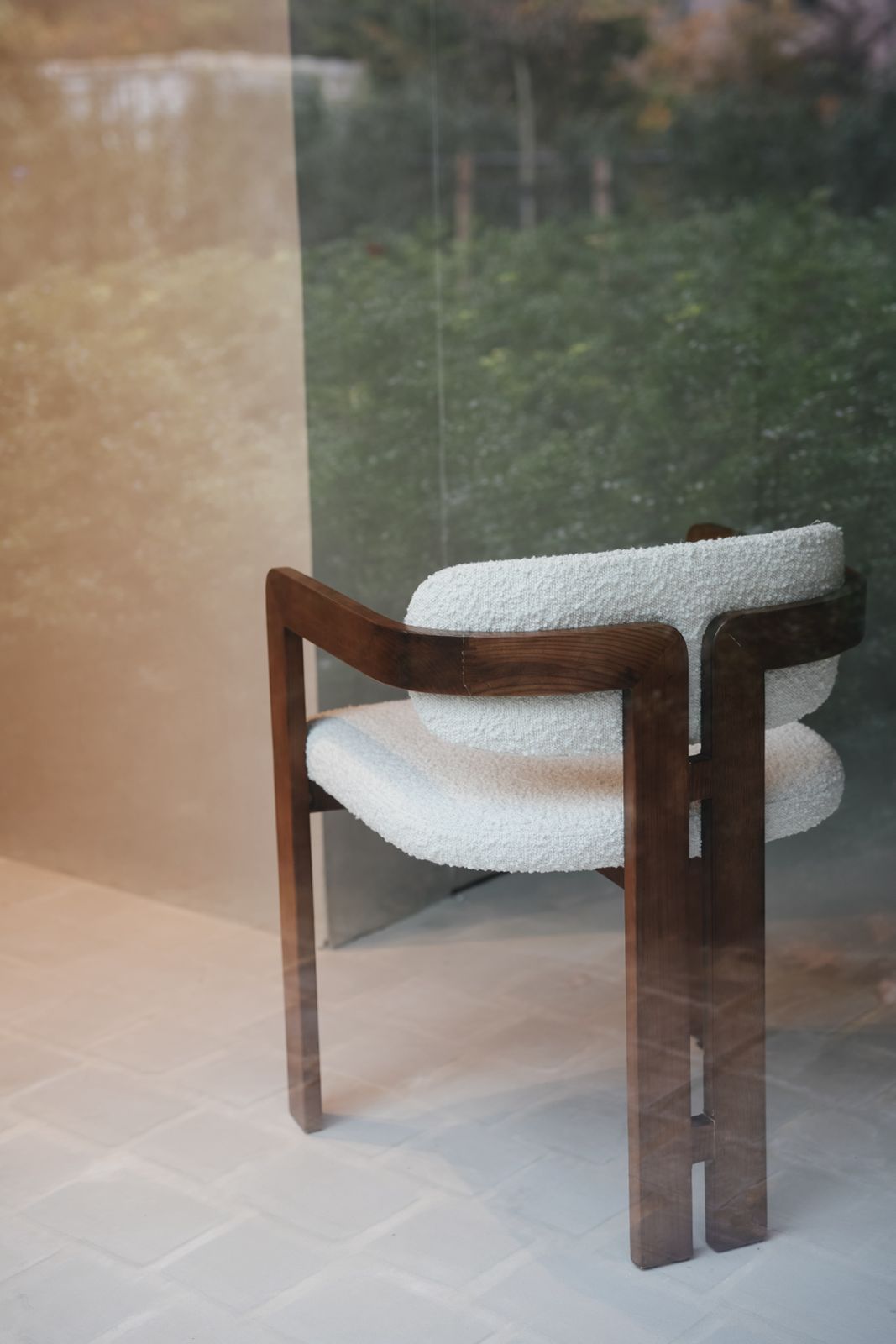 Back view of Mila dining chair by Mowelli in ash wood and white bouclé fabric, seen through window. 