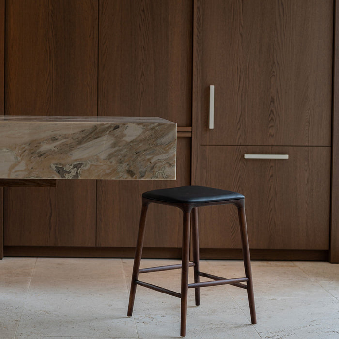 Mowelli Nora counter stool in brown walnut wood with premium vegan leather upholstered seat and minimalist backless design.
