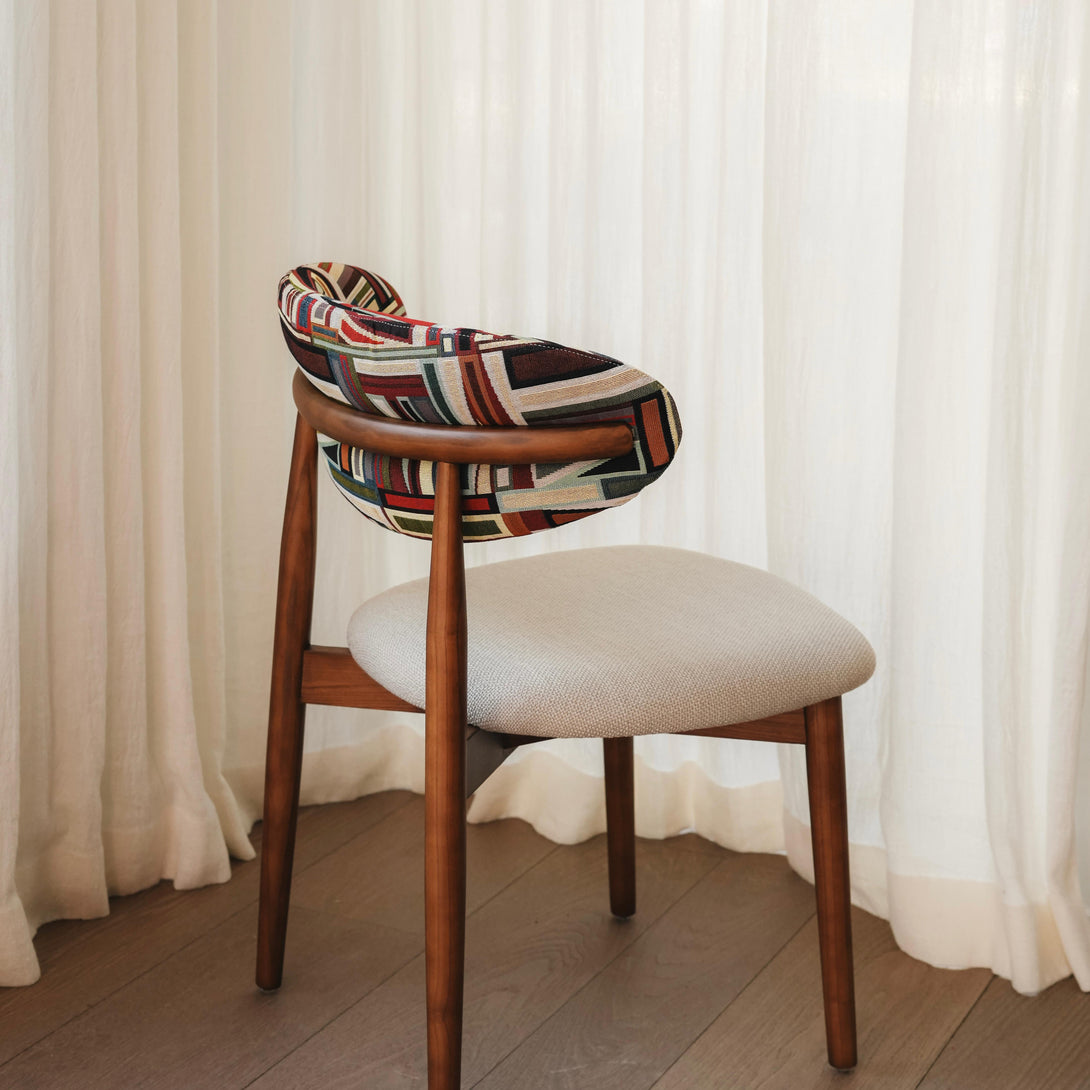 A modern dining chair with a neutral colored seat and a geometric multicolored backrest, set against a white curtain backdrop.