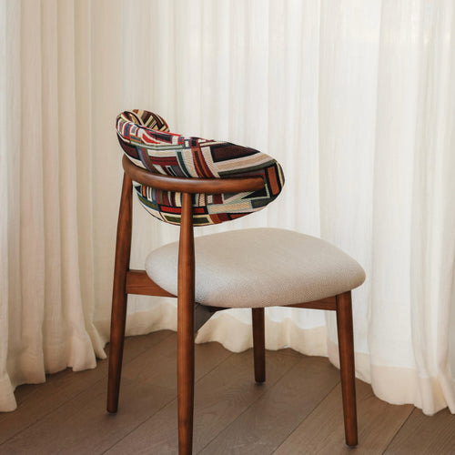 A modern dining chair with a neutral colored seat and a geometric multicolored backrest, set against a white curtain backdrop.