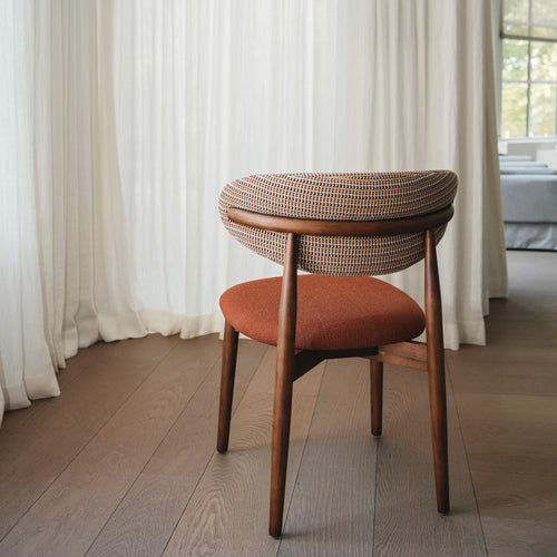 A modern dining chair with a terracotta-colored seat and a backrest featuring a mix of cheerful colors, placed in a room with white curtains and wooden flooring.