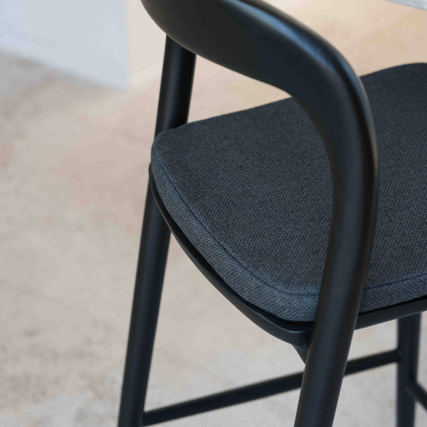 A close-up image of a charcoal black fabric cushion on a Nora bar stool, showcasing the texture and color of the seat cushion.
