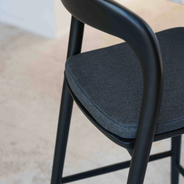 A close-up image of a charcoal black fabric cushion on a Nora bar stool, showcasing the texture and color of the seat cushion.