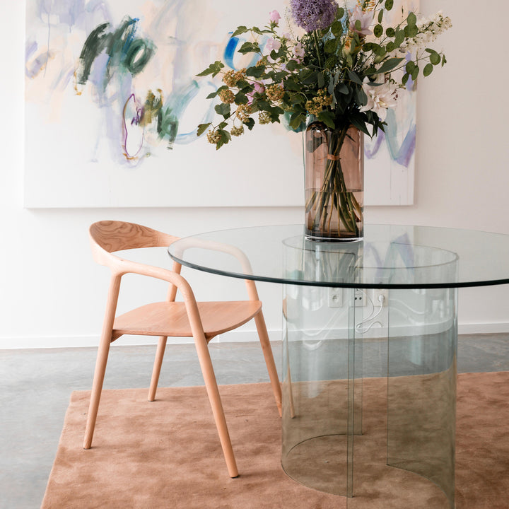 Ash wood Nora dining chair by Mowelli next to a round glass table in a light-filled interior.
