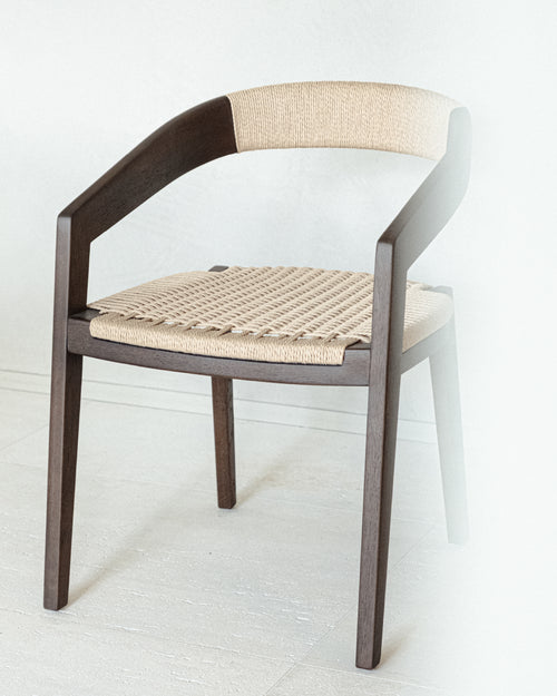 A dining armchair of solid matt dark brown ash wood, with a floating backrest that curves into the armrests. The seat and back are woven in natural paper cord.