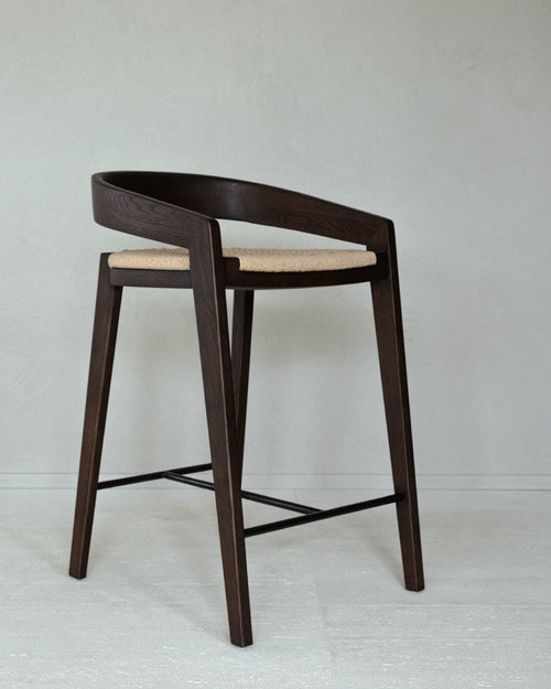 A kitchen stool of solid matt dark brown ash wood, with a floating backrest that curves into the armrests. The seat is woven in natural paper cord.