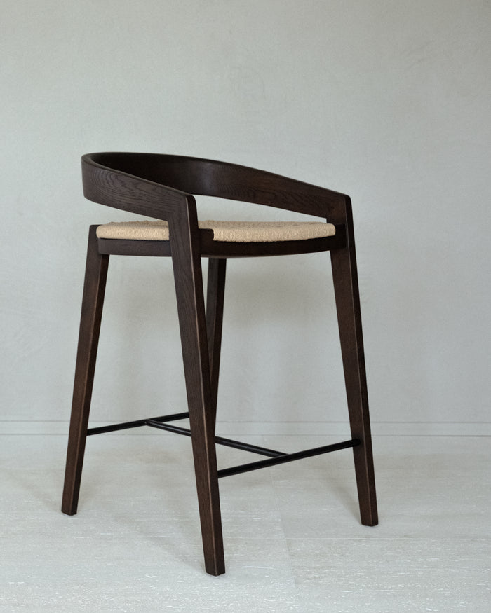 A kitchen stool of solid matt dark brown ash wood, with a floating backrest that curves into the armrests. The seat is woven in natural paper cord.