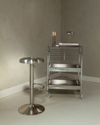 Gigi bar stool in brushed stainless steel next to a metal trolley in a minimalist interior