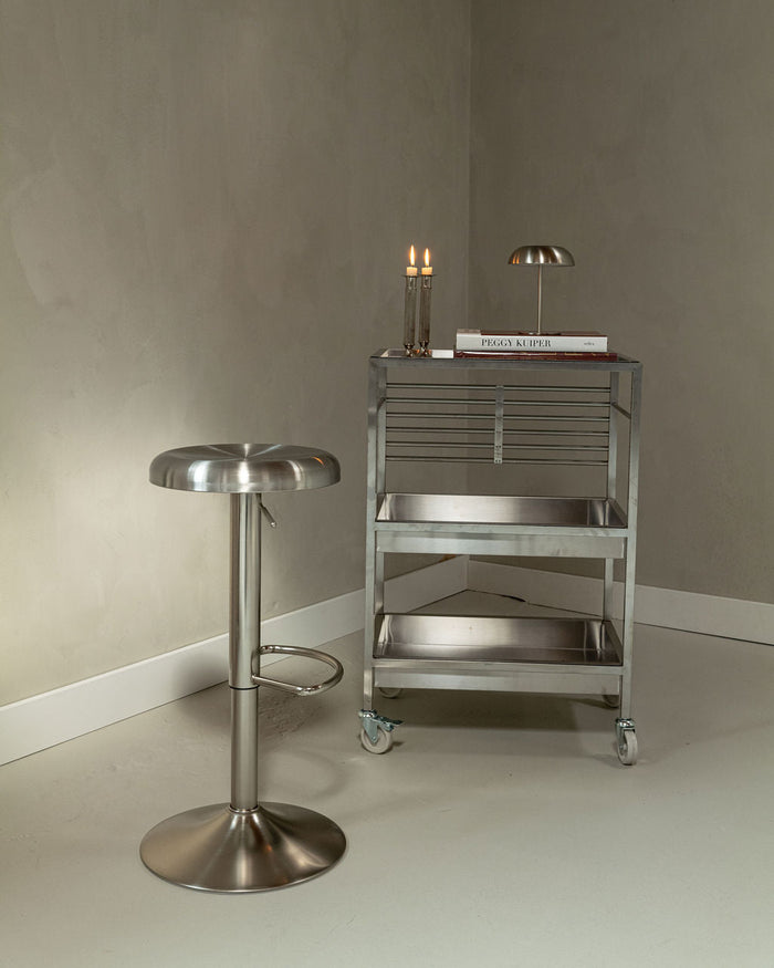 Gigi bar stool in brushed stainless steel next to a metal trolley in a minimalist interior