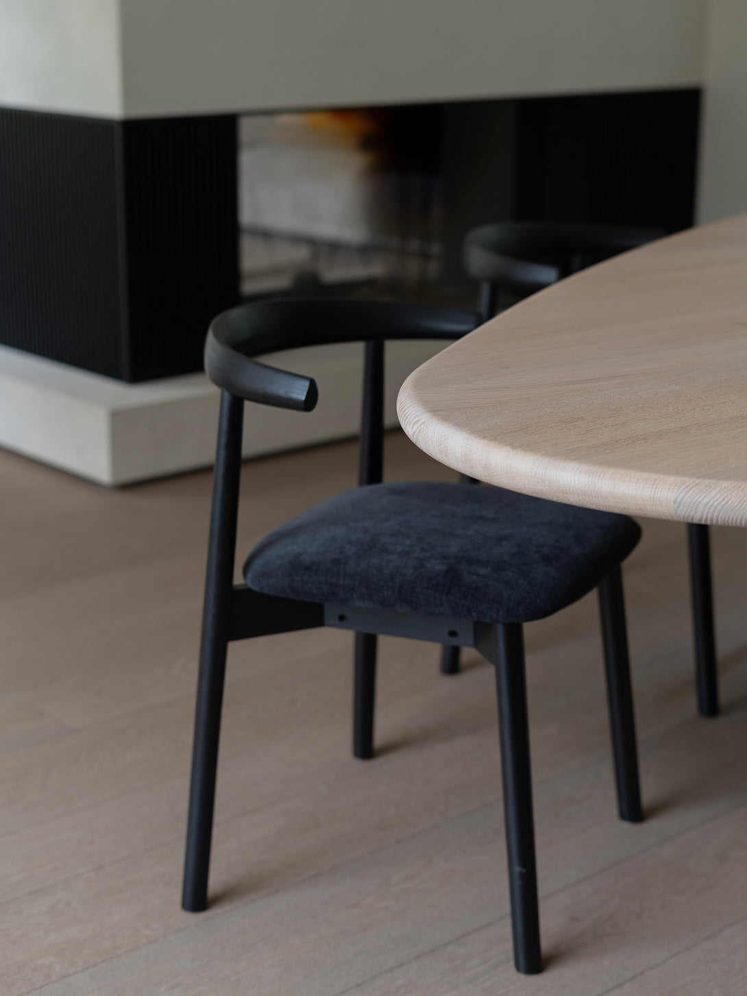 Ella dining chair by Mowelli with a upholstered seat and wooden backrest, all black, around a table, fireplace in the back. 