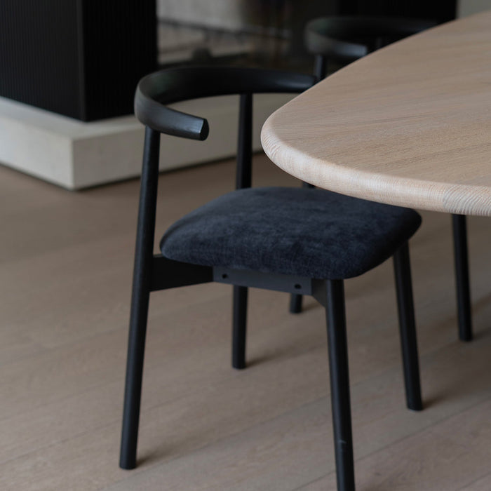 Ella dining chair by Mowelli with a upholstered seat and wooden backrest, all black, around a table, fireplace in the back. 